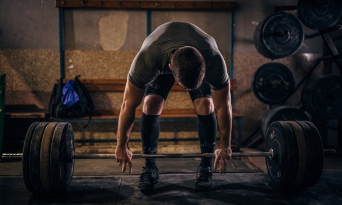 A personal trainer in the area is exercising at a gym. The photo shows a man about 30 years old in a dark room lifting a barbell