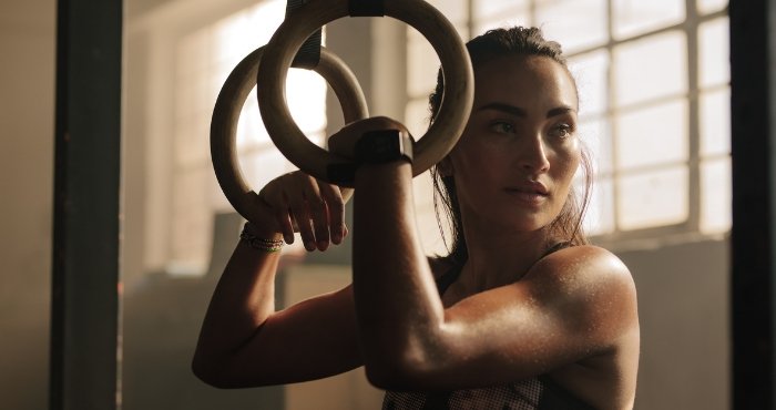 The woman trains at a gym taught by a personal trainer in her area. The woman stands illuminated by the sunset and holds on to rings hanging from the ceiling